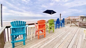 A wooden deck with colorful Adirondack chairs and a black umbrella, overlooking a beach.