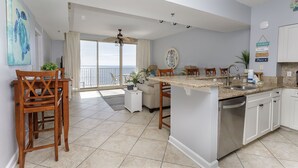 A spacious kitchen with a large island, a dining area with chairs, and a view of the ocean through the sliding glass doors. - Shores of Panama 2BR Unit 1911! 19th Floor Gulf Views! Luxury Furnishings! (Panama City Beach)