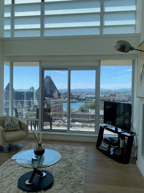 A spacious living room with a large window offering a city view, a round glass table, a sofa, and a television. - Spacious Splendour-Panoramic Views from 12th Floor Luxury Condo, AC, Pool and Parking (Victoria)