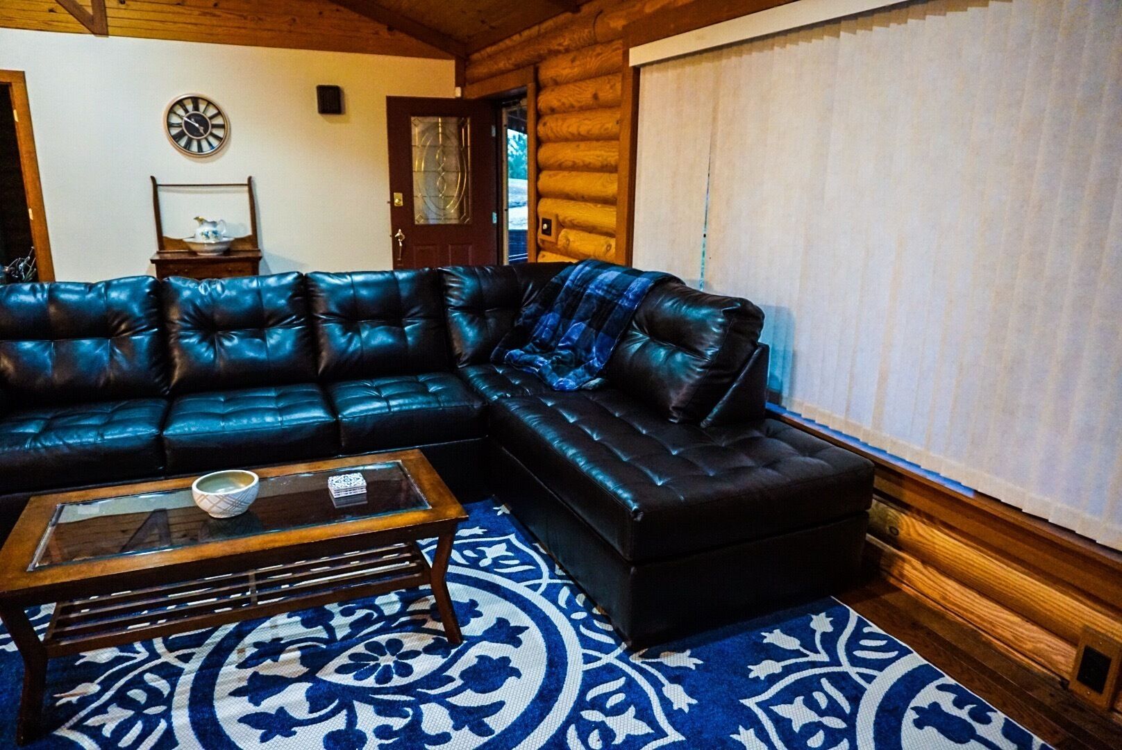 A log cabin interior with a black leather sectional sofa, a wooden coffee table, and a patterned rug.