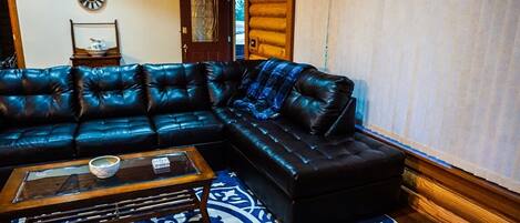 A log cabin interior with a black leather sectional sofa, a wooden coffee table, and a patterned rug.
