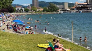 A crowded lakeside beach with people swimming and sunbathing, surrounded by buildings and hills. - Premiere Beach House, Family Friendly, West Lakeshore Drive (coeur d'alene)