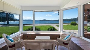 A sunroom with large windows overlooking a lake, wicker furniture, and a view of the water and greenery. - Premiere Beach House, Family Friendly, West Lakeshore Drive (coeur d'alene)
