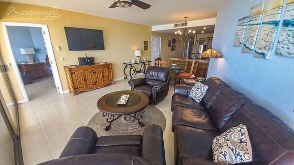 A living room with a brown leather sofa, a coffee table, and a TV. There's a dining area with a chandelier and a table. - Sonoran Spa W603 - 2 BR Next to the Pool, High Speed Wi-Fi - Beachfront Upper Floor Luxury Condo (Puerto Peñasco)