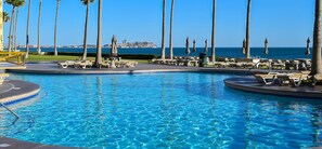 Zona de piscina con sillones de descanso y palmeras, vista despejada del océano y una isla al fondo.