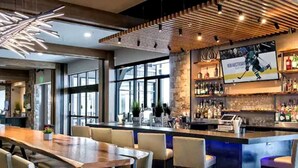 A bar area with a long wooden table and several bar stools, a stone accent wall, and a large TV screen displaying a hockey player. - Luxury Ski-in/Ski-out residence (Breckenridge)