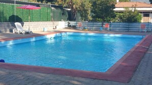 A rectangular swimming pool with a red border, surrounded by a brick wall and a green fence. There are lounge chairs and a red umbrella near the pool. - Camping Blumare Village (Furnari)