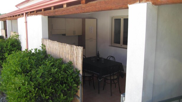 A covered outdoor dining area with a table and chairs, a wooden pergola, and a window overlooking a garden. - Camping Blumare Village (Furnari)