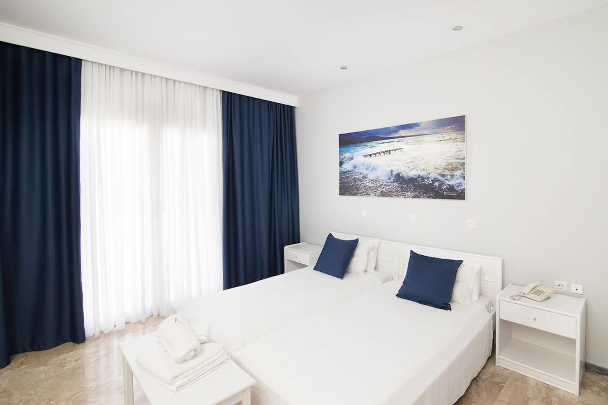 A hotel room with a large bed, white bedding, two pillows, a nightstand with a phone, and a painting of a coastal scene.
