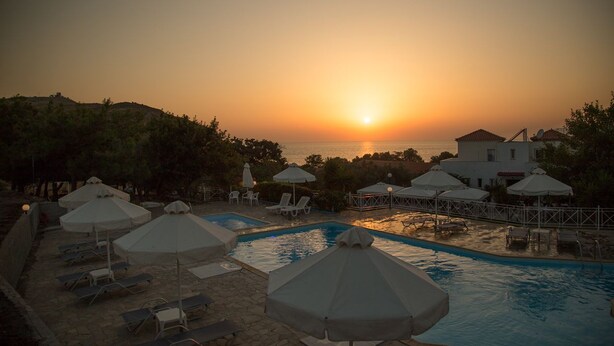 Zona de piscina con sombrillas y sillas de descanso, con vista a la puesta de sol sobre el mar.