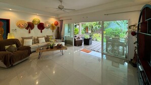 Amplia sala de estar con sofá, mesa de centro y ventiladores decorativos. Unos ventanales corredizos dan a un patio exterior con zona de comedor.