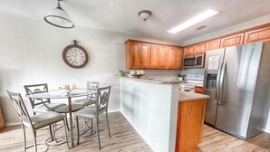 Una cocina con armarios de madera, un refrigerador de acero inoxidable, un microondas y un comedor con mesa de vidrio y sillas de metal.