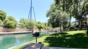 Ein Picknickbereich am Flussufer mit Holztisch und Grill, umgeben von Bäumen und einer Steinmauer.
