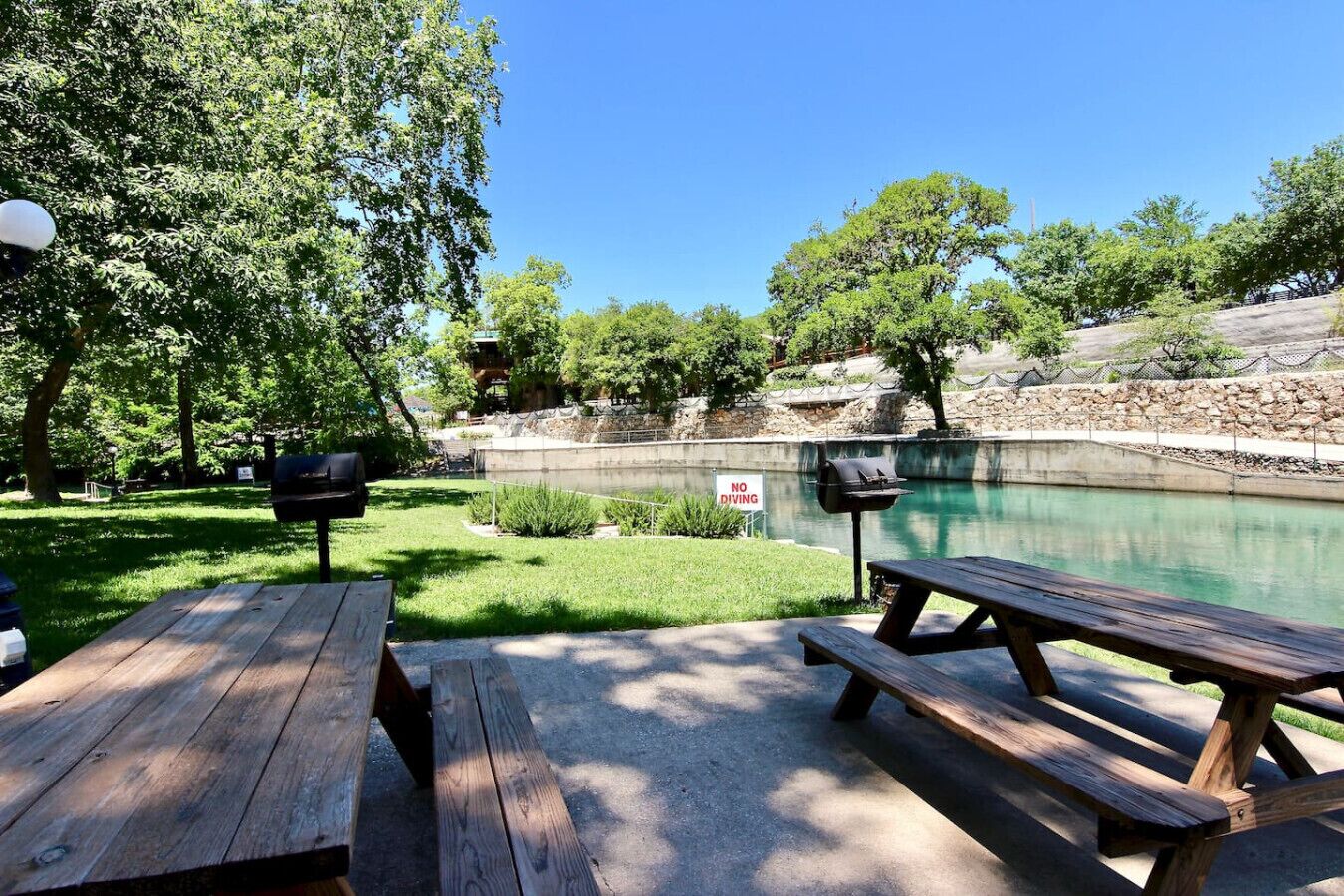 A picnic area with wooden benches and a grill, overlooking a calm pond with a 'NO DIVING' sign.