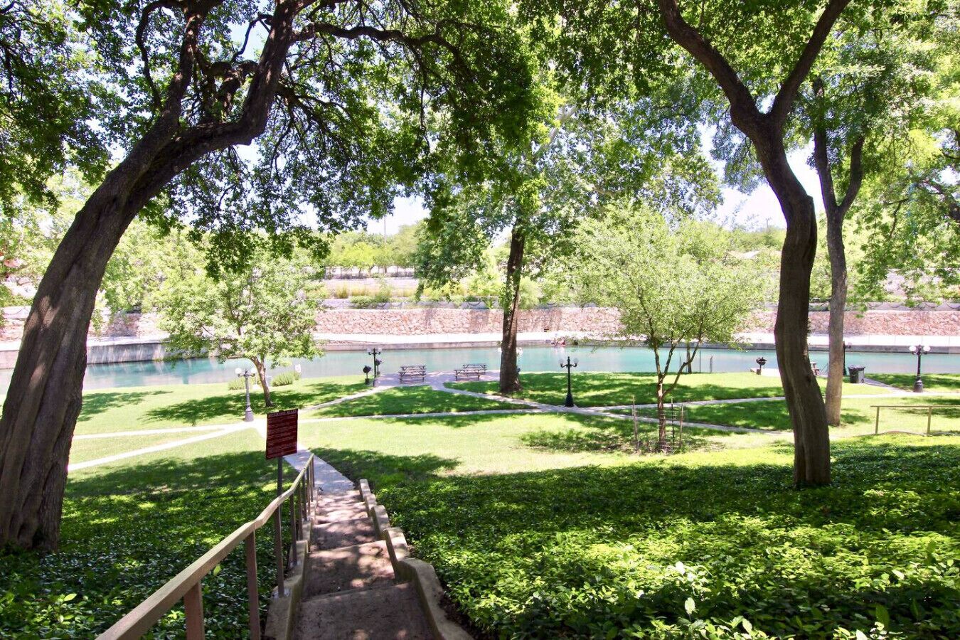 A park with a pathway leading to a body of water, surrounded by trees and benches.