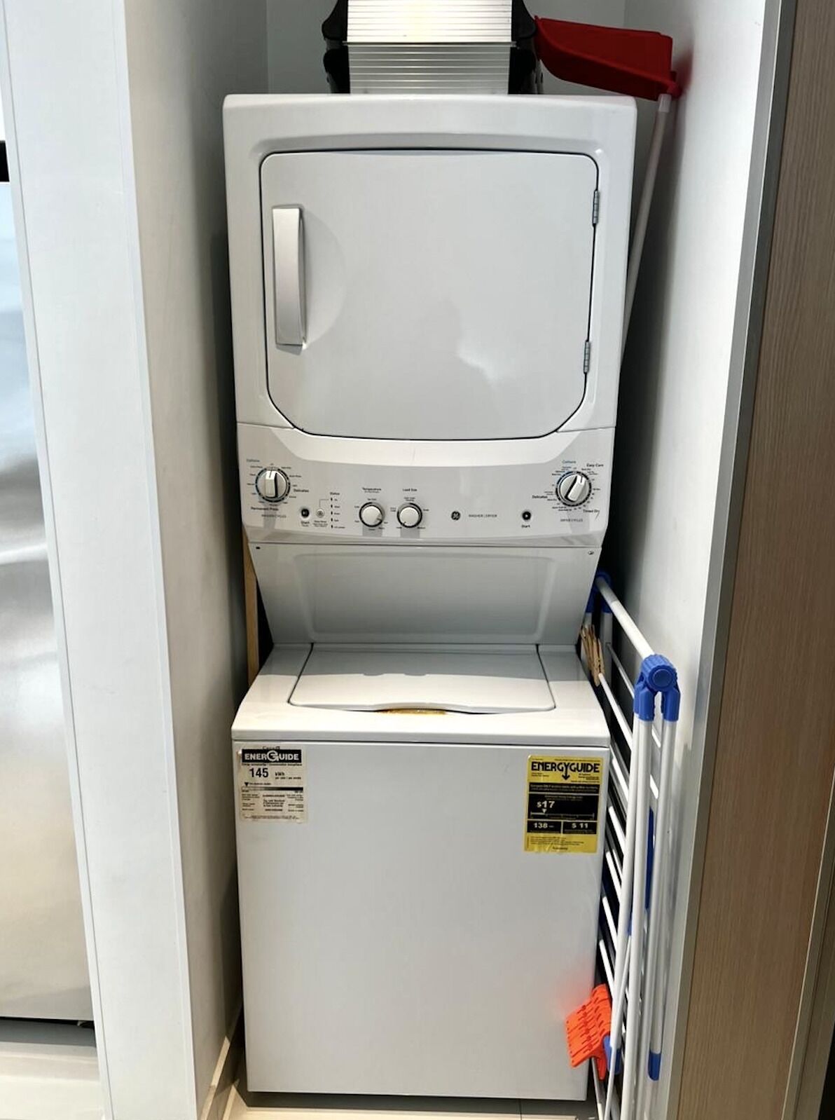 Un appareil compact combinant lave-linge et sèche-linge, doté d’un panneau de commande, d’une étiquette indiquant la consommation énergétique et d’un tuyau rouge et bleu.