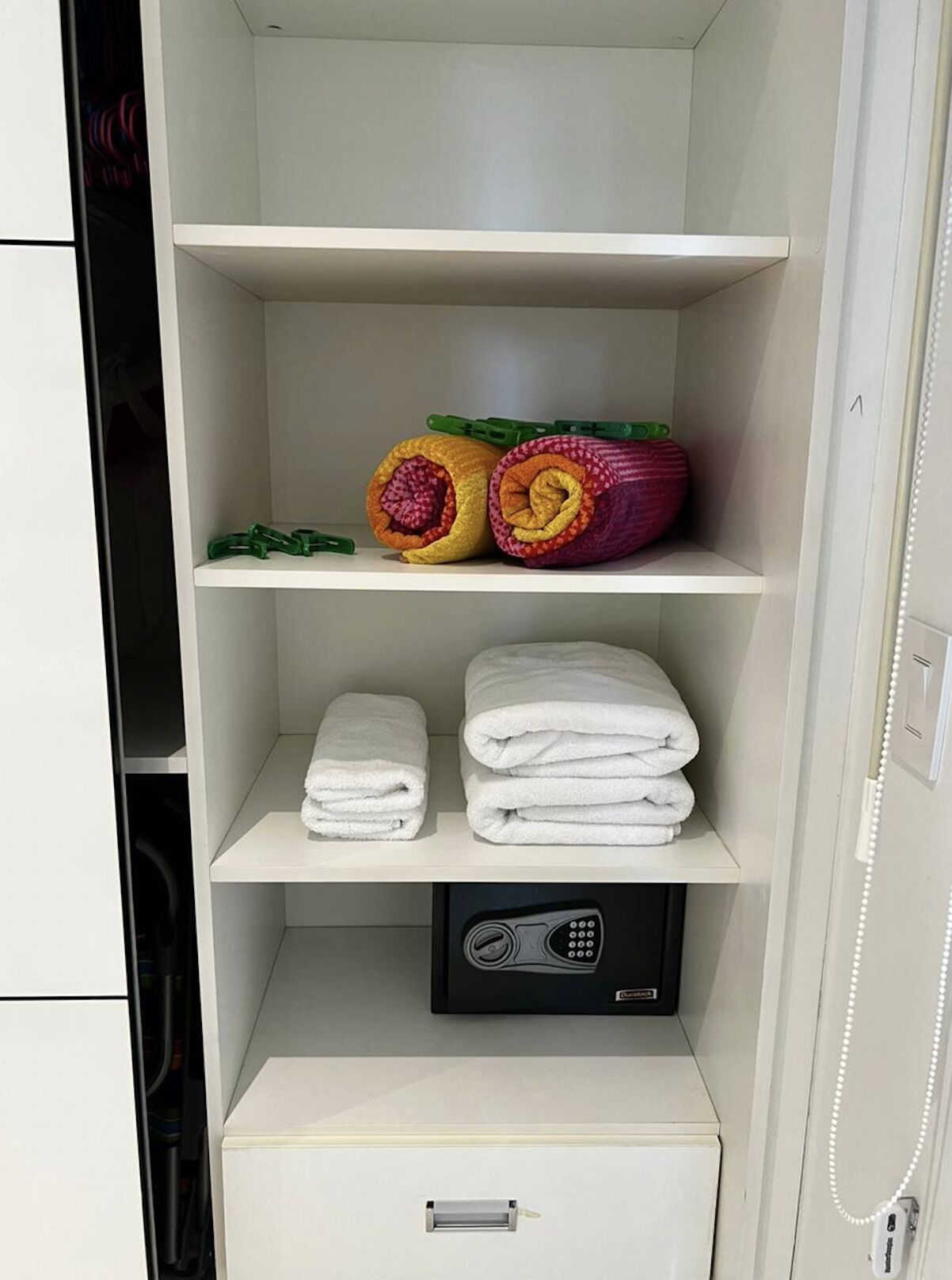 Habitación de hotel con un armario blanco que contiene toallas blancas ordenadas, un juego de toallas enrolladas de colores y un dispositivo electrónico negro en la repisa inferior.