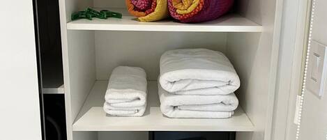 Habitación de hotel con un armario blanco que contiene toallas blancas ordenadas, un juego de toallas enrolladas de colores y un dispositivo electrónico negro en la repisa inferior.