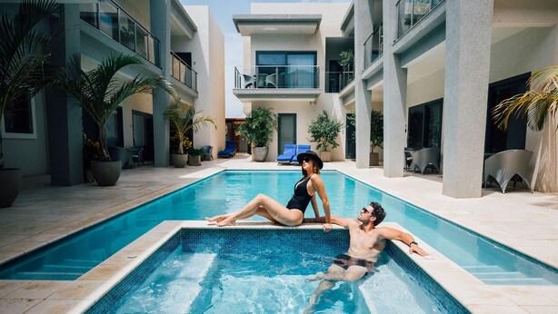 A modern indoor pool area with a couple relaxing by the pool, surrounded by potted plants and lounge chairs.