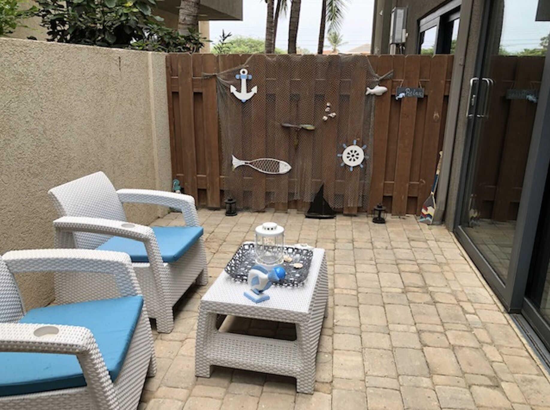 Un patio con mobiliario de mimbre y una pequeña mesa, una valla de madera decorada con motivos náuticos, y un ventanal de vidrio que da a una zona cubierta.