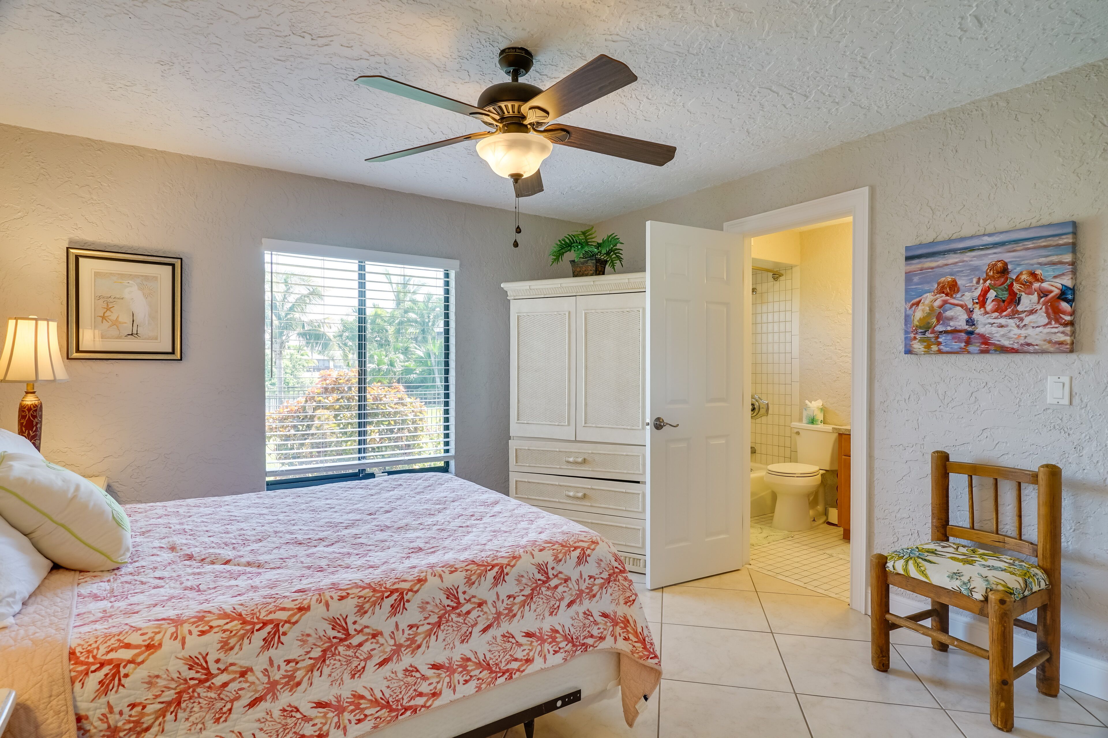 A bedroom with a bed, a ceiling fan, a window with blinds, a door leading to a bathroom, and a chair.