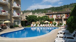 A swimming pool with lounge chairs and umbrellas, surrounded by buildings with balconies and flower boxes. - Hotel Romeo (Torri del Benaco)