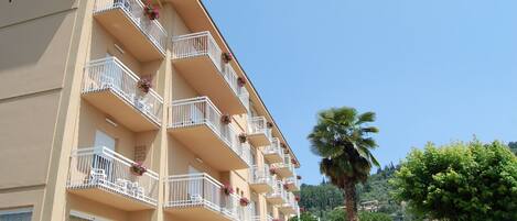 Un hotel de varios pisos con balcones decorados con flores, cielo despejado y abundante vegetación al fondo.