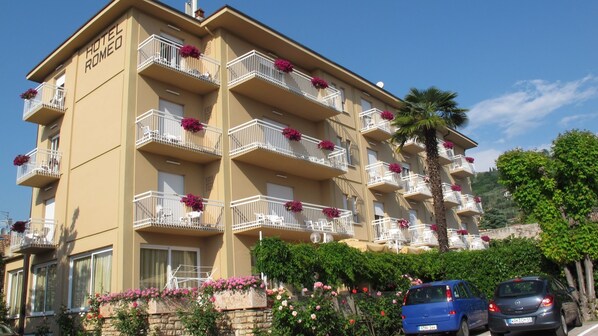 Un hôtel à plusieurs étages, doté de balcons ornés de fleurs, d’un parking rempli de voitures et entouré d’une végétation luxuriante.