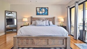 Une chambre avec un lit en bois, deux lampes de chevet, un ventilateur de plafond, une grande fenêtre avec des rideaux et une œuvre d’art abstraite accrochée au mur.