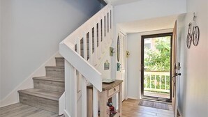 Un escalier avec des marches en bois et une rampe blanche, une petite console en bois et une porte donnant sur un balcon agrémenté de plantes.