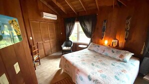 A wooden cabin bedroom with a bed, a small table, a chair, and a window with curtains. - Monticello, Desert Rose beachfront cottage,Tiki Hut & Generator,10% Off 7+nights (governors harbour)