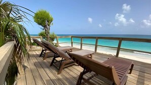 A wooden deck with lounge chairs and a table overlooking a beach with clear turquoise water. - Monticello, Desert Rose beachfront cottage,Tiki Hut & Generator,10% Off 7+nights (governors harbour)