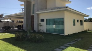 A modern two-story house with a flat roof, large glass sliding doors, and a small garden area with stepping stones leading to the entrance. - Modern house in Barra do Jacuípe, 180m from the beach. (Camaçari)