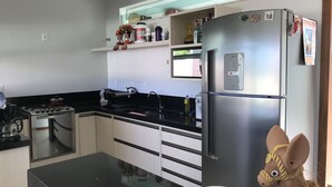 A modern kitchen with a stainless steel refrigerator, a stove, and a sink. There are shelves above the counter holding various items. - Modern house in Barra do Jacuípe, 180m from the beach. (Camaçari)