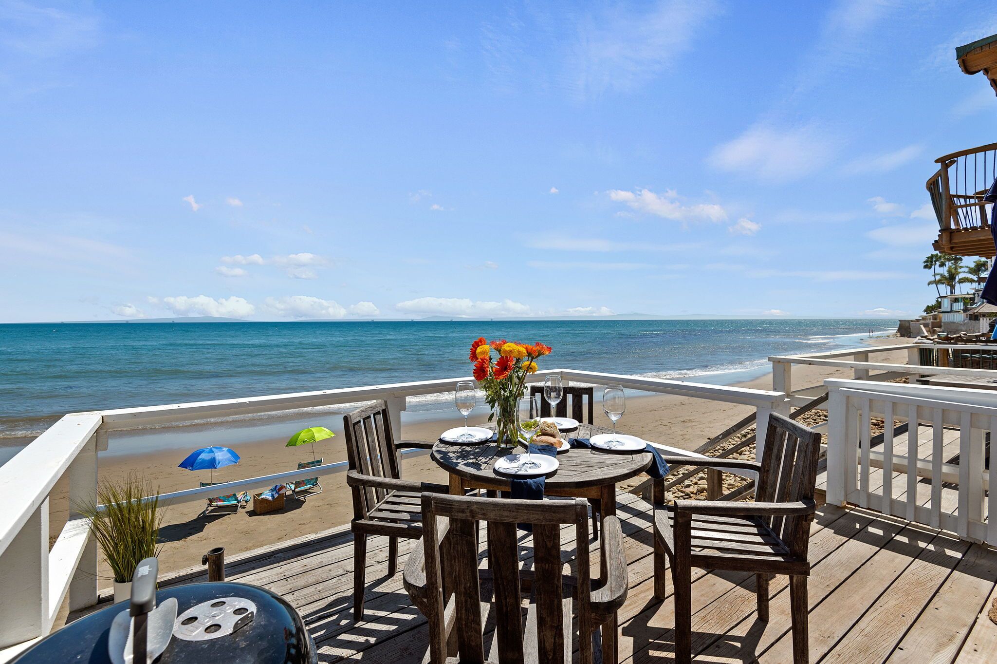A wooden deck with a table set for dining, overlooking a beach with umbrellas and lounge chairs.