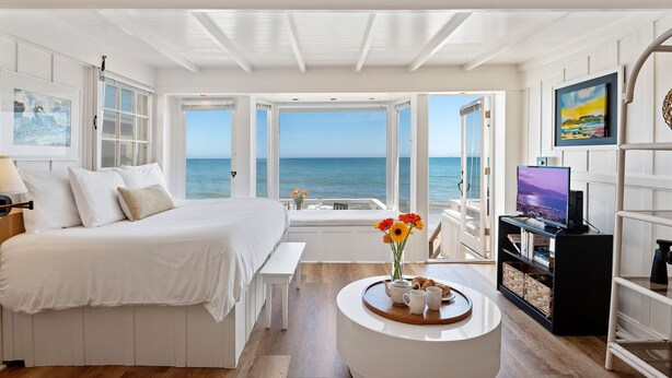 A bedroom with a large bed, a bedside table, a round coffee table with a vase of flowers, and a TV on a stand. - Romantic Oceanfront Studio With Private Deck | Right On The Sand! (Santa Barbara)