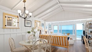 A spacious room with a large window overlooking the ocean, a wooden ceiling, and a dining area with wicker furniture. - Romantic Oceanfront Studio With Private Deck | Right On The Sand! (Santa Barbara)