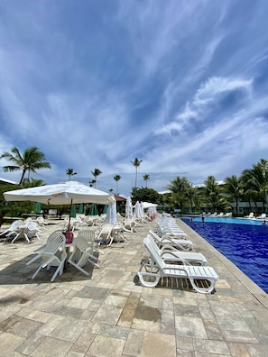 A poolside area with white lounge chairs and umbrellas, a swimming pool, and palm trees under a cloudy sky.