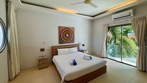 A modern bedroom with a large bed, a ceiling fan, an air conditioning unit, a bedside table with a lamp, and a window with curtains.