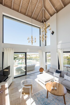 Relax in this stylish living area with a high ceiling, featuring a large chandelier, comfortable sofa, and a TV on a stand.