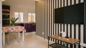 A modern kitchen with a dining area, a flat-screen TV mounted on the wall, and a table with a vase of flowers.