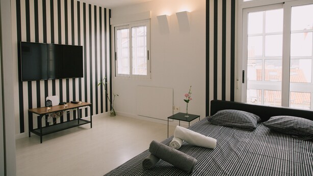 A modern bedroom with a black and white striped wall, a flat-screen TV, a bed with patterned bedding, and a window with a city view.