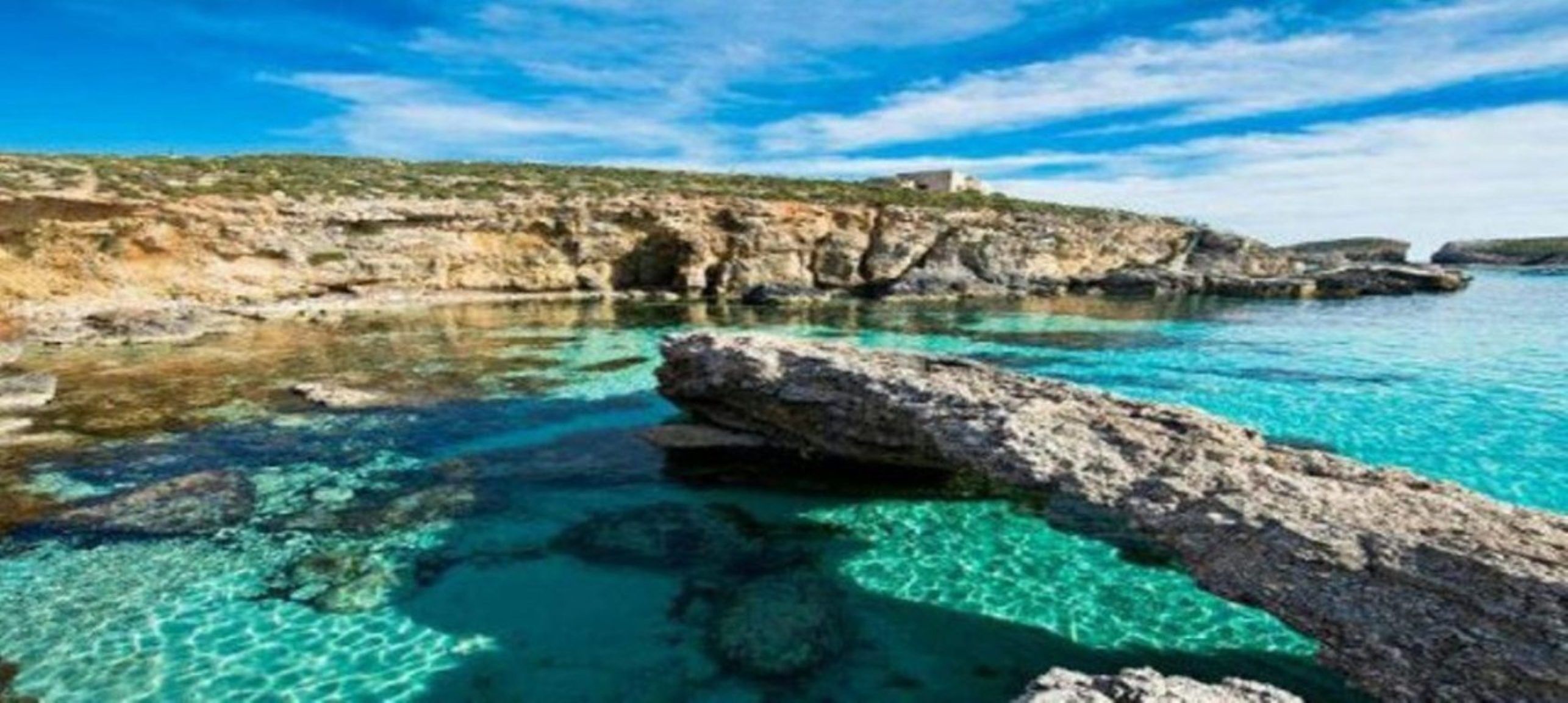 A coastal landscape with clear turquoise waters, rocky formations, and a distant building on a cliff.