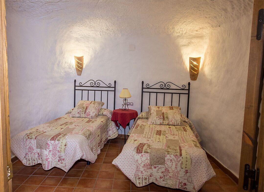 A room with two single beds, each with a patterned bedspread, a small table with a lamp, and wall-mounted lights.