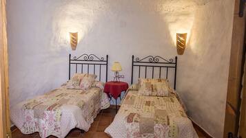 A room with two single beds, each with a patterned bedspread, a small table with a lamp, and wall-mounted lights.