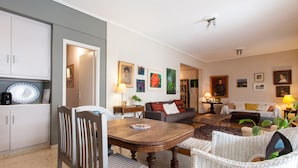 A cozy living room with a dining table, chairs, a sofa, and a coffee table. There are framed pictures on the wall and a lamp on a side table.