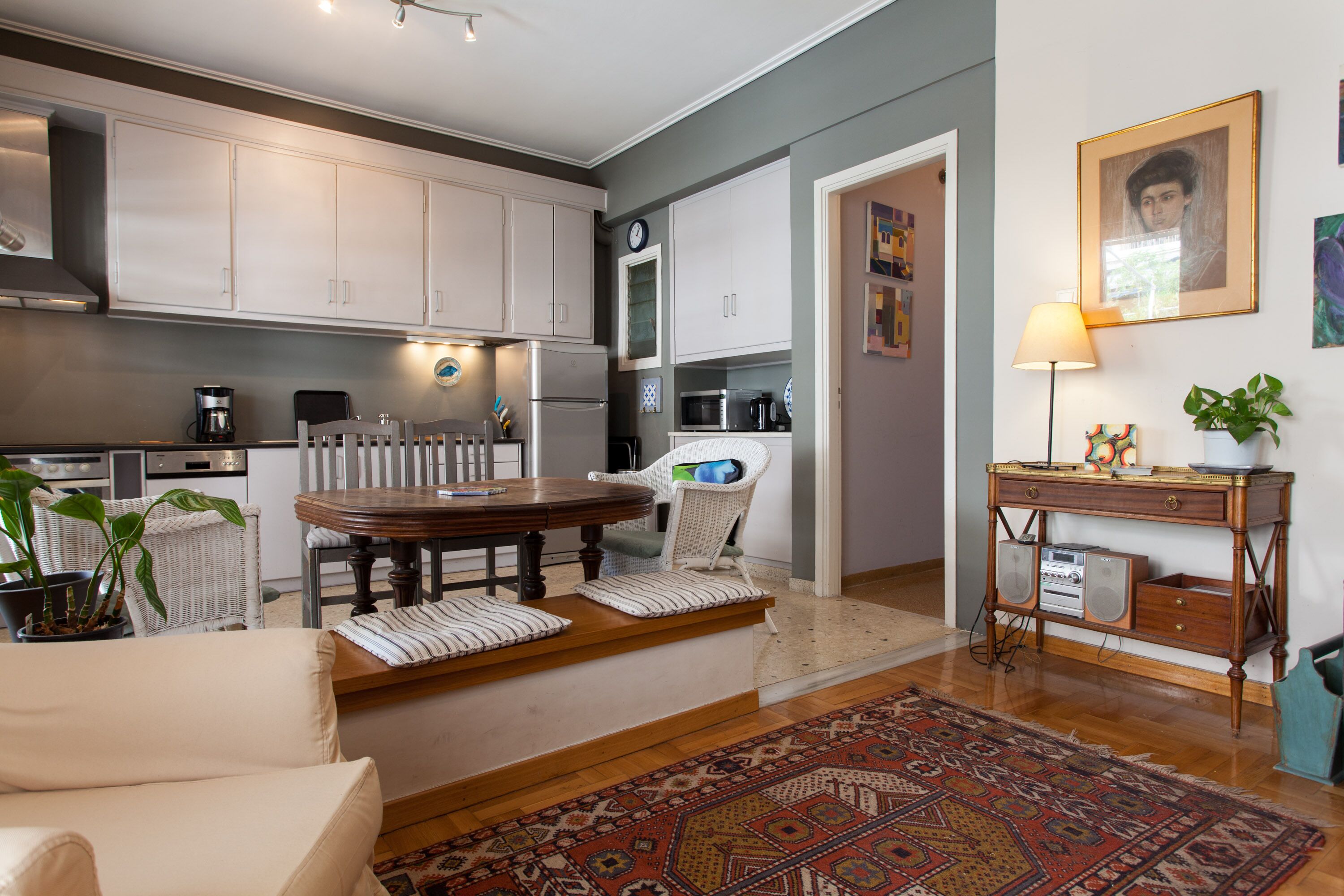Eine moderne Küche mit weißen Schränken, ein Essbereich mit einem Holztisch und Stühlen sowie ein Wohnzimmer mit einer Couch und einem gemusterten Teppich.