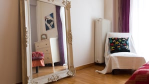 A bedroom with a large ornate mirror, a white radiator, a bed with a colorful pillow, and a dresser with a pink towel.