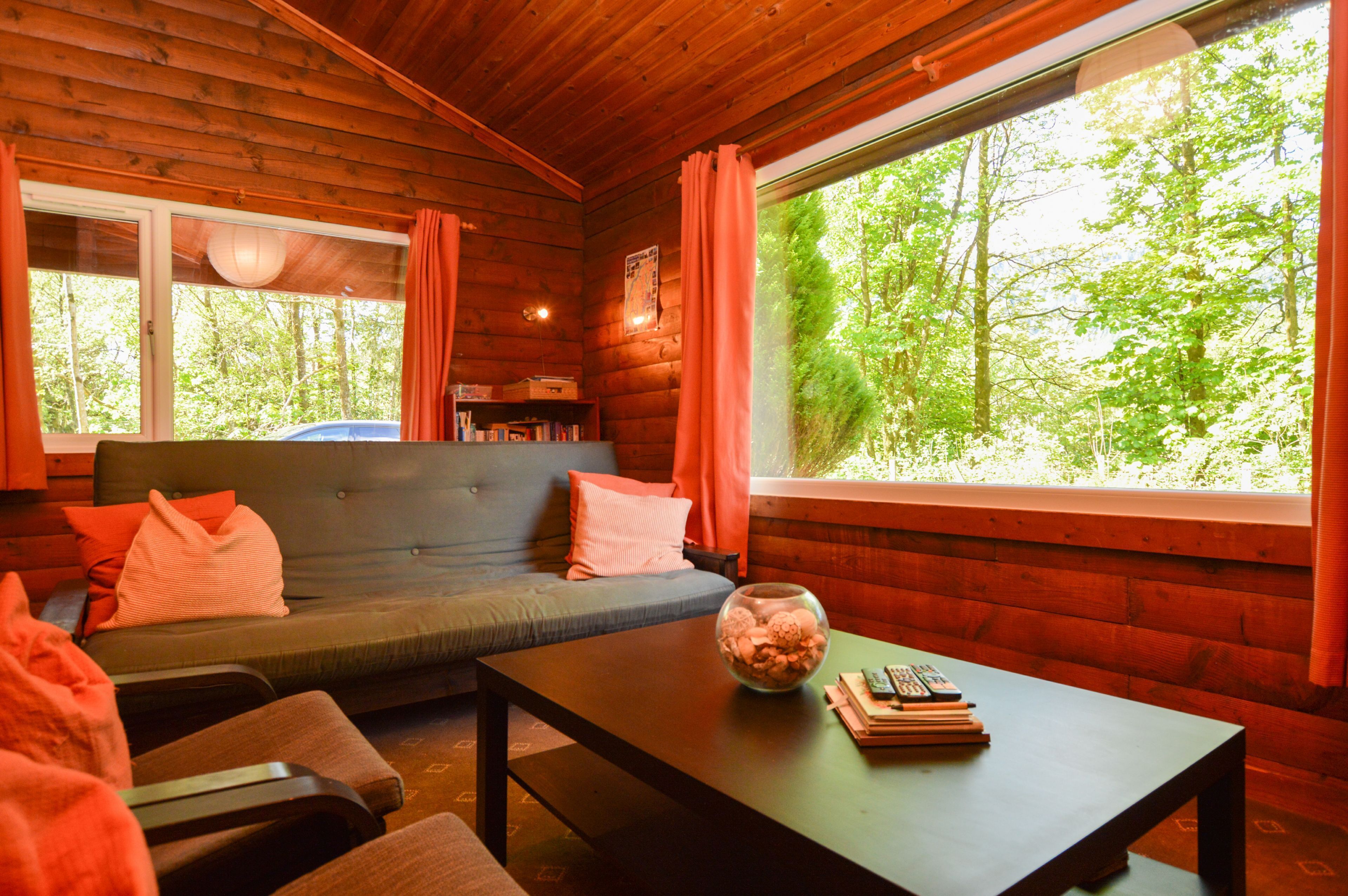 A cozy cabin interior with a sofa, a coffee table, and a large window offering a view of a forest.