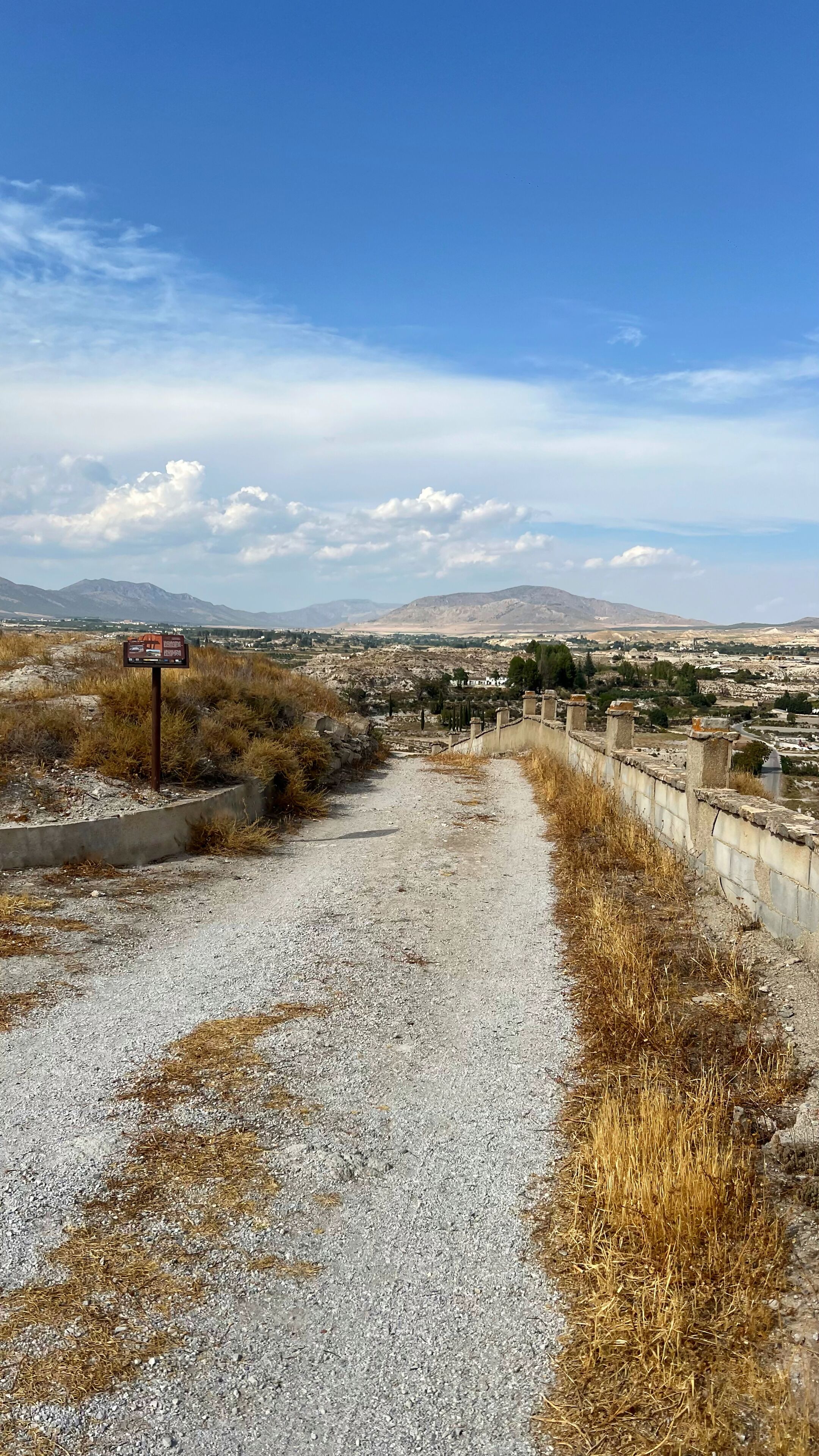 Un sendero de grava que conduce a un sitio histórico con un cartel, rodeado de hierba seca y un cielo despejado.